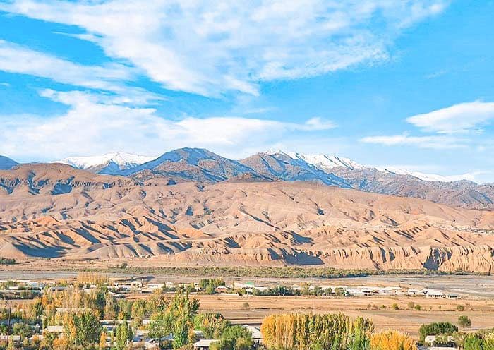 Arid mountain landscape with snow-capped peaks, dry foothills, and a small settlement with autumn trees in the foreground under a blue sky.