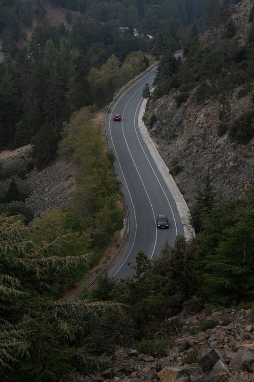Winding two-lane asphalt road through forested hills with two cars driving, viewed from above.