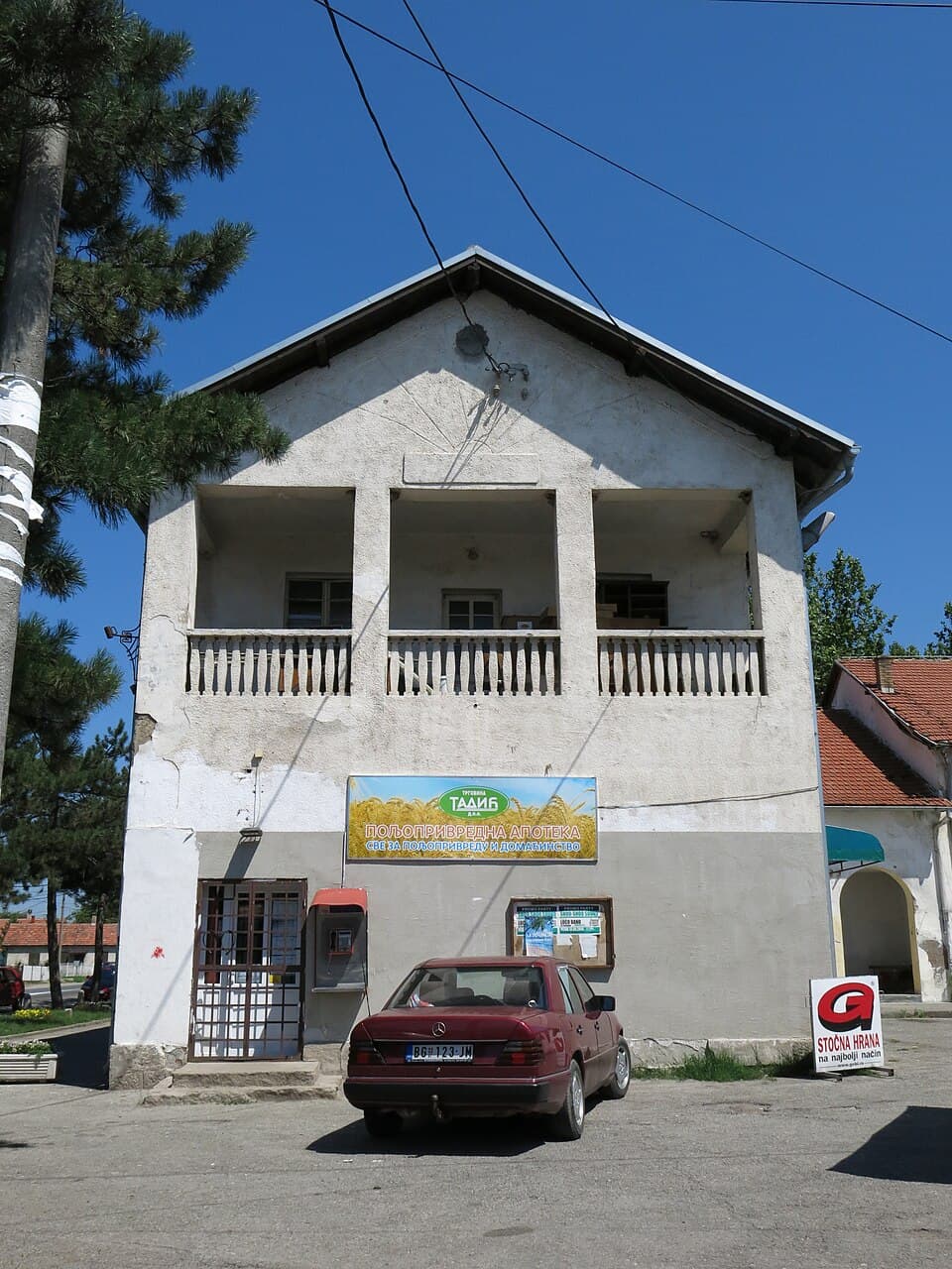 A white two-story building with a balcony and Cyrillic signage stands under a blue sky, with a red car parked in front.