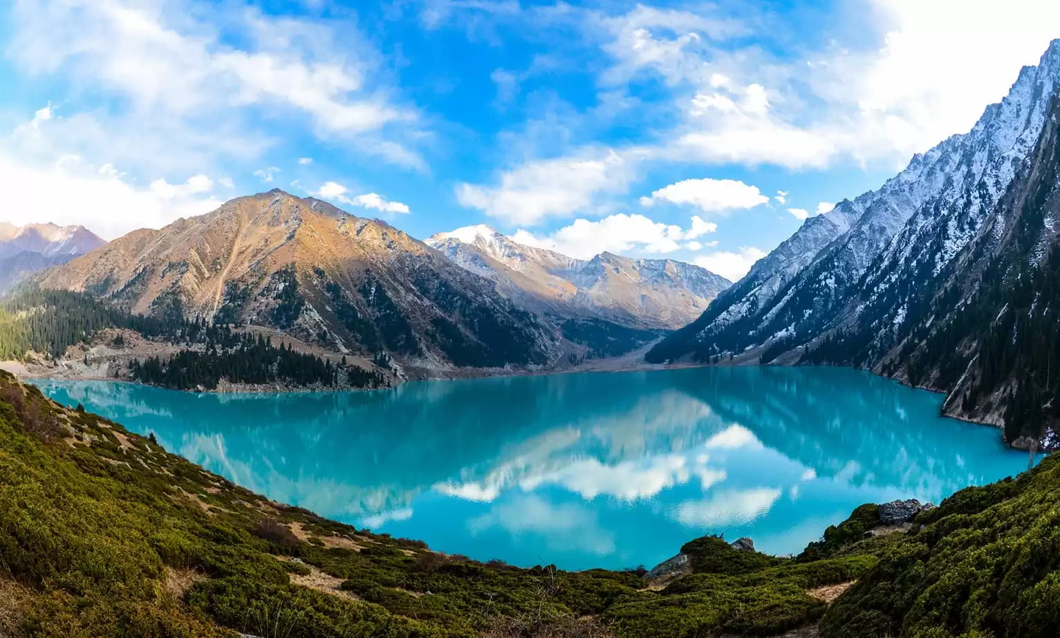 Turquoise alpine lake surrounded by snow-capped mountains and green slopes in the Bishkul area of Kazakhstan under a partly cloudy sky.
