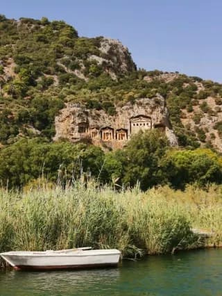 Lycian Rock Tombs carved into a hillside overlook the Dalyan River, with a small boat moored near reeds in the foreground.