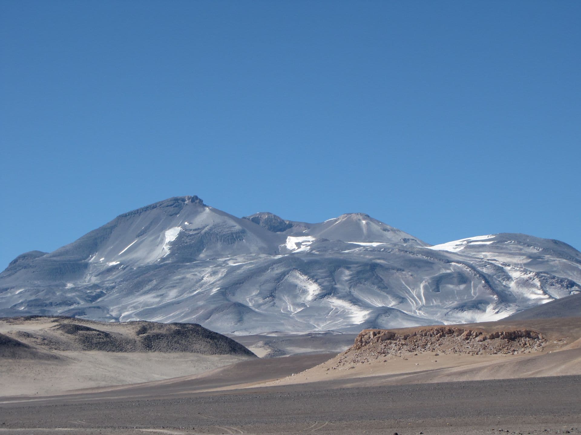 Arid desert landscape near Provincia de Copiapó, Chile, with a gravel road in the foreground leading toward snow-dusted mountains under a clear blue sky.