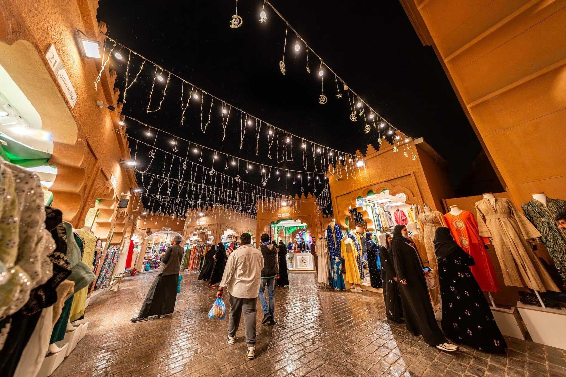 Nighttime market alley in the Jizan region with hanging lights, stalls, and shoppers on wet pavement