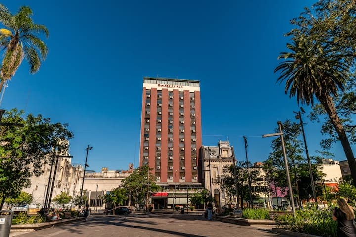 A sunny street scene in Simoca featuring a tall red brick building with a white top, flanked by palm trees and a clear blue sky.