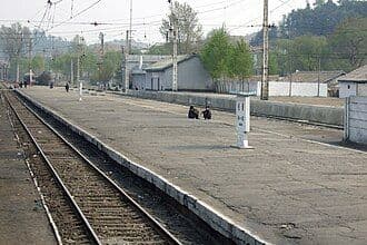 Wide shot of a railway station platform with tracks, overhead wires, and a few distant figures.