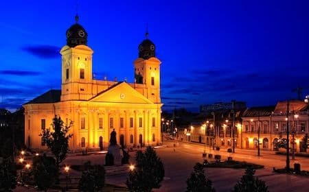 Night view of the illuminated Great Church in Debrecen, featuring twin towers and a statue in the foreground against a deep blue sky.