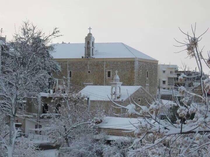 Snow-covered stone church with bell tower and cross, surrounded by frosted trees and village buildings under an overcast sky.