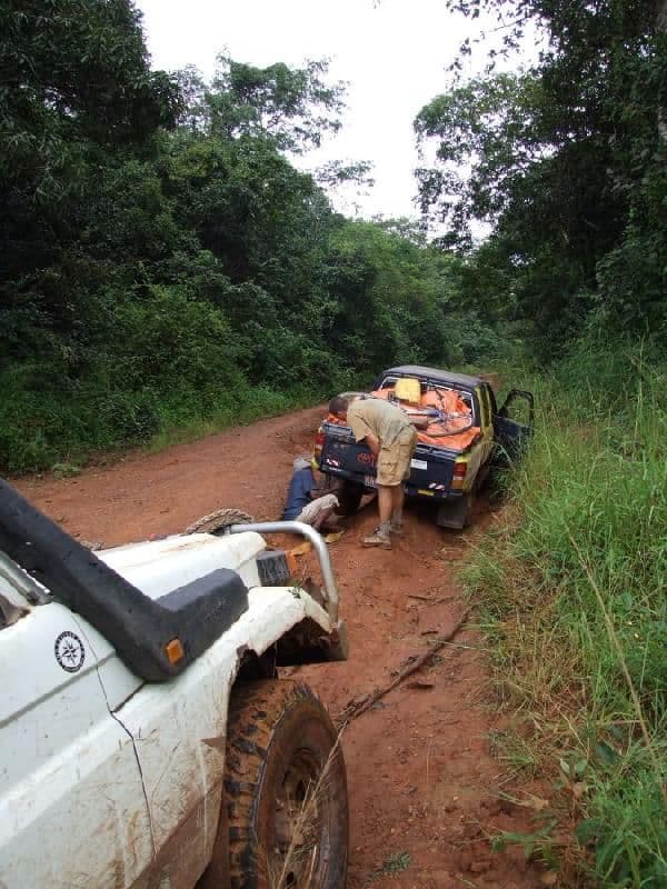 Two pickup trucks on a red dirt road in dense forest; one is stuck while men work to free it.