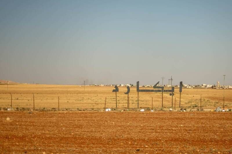 Sunlit rural landscape with golden fields, a large Arabic sign, and distant buildings under a clear blue sky.