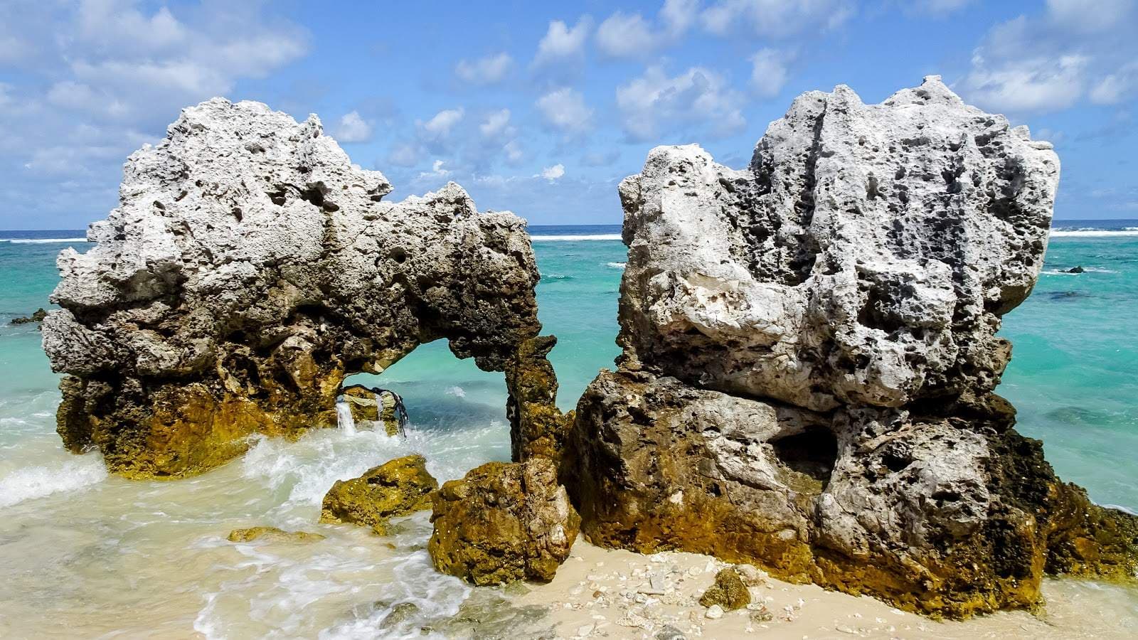 Coastal shoreline with large white rock formations, turquoise water, and sandy beach under a partly cloudy sky.
