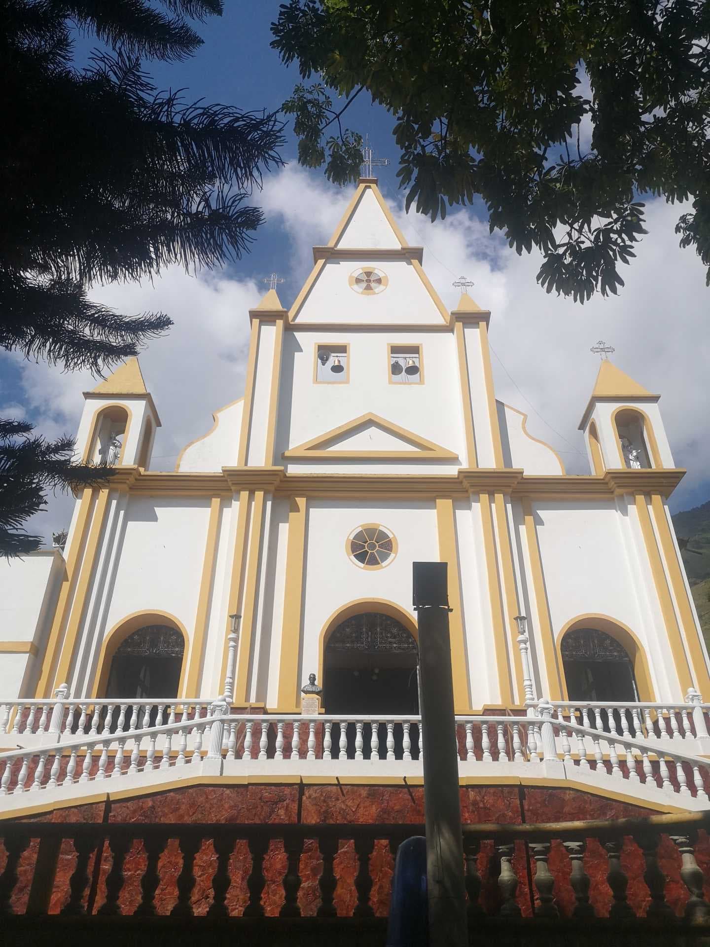 White and yellow church facade in Liborina with twin bell towers, arched entrances, and a balustraded staircase, framed by tree branches.