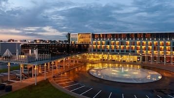 Evening view of a modern hotel exterior with illuminated windows, a central courtyard pool, and a parking area under a cloudy sky.