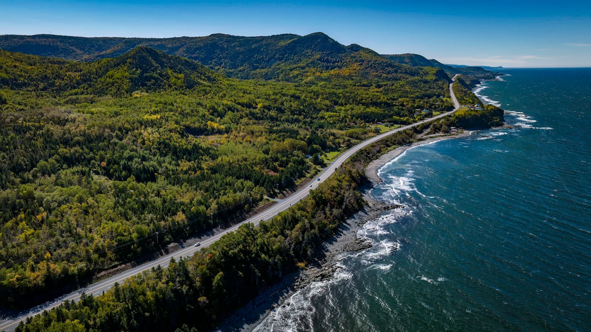 Aerial view of a coastal highway winding through forested mountains beside a rocky shoreline and blue ocean