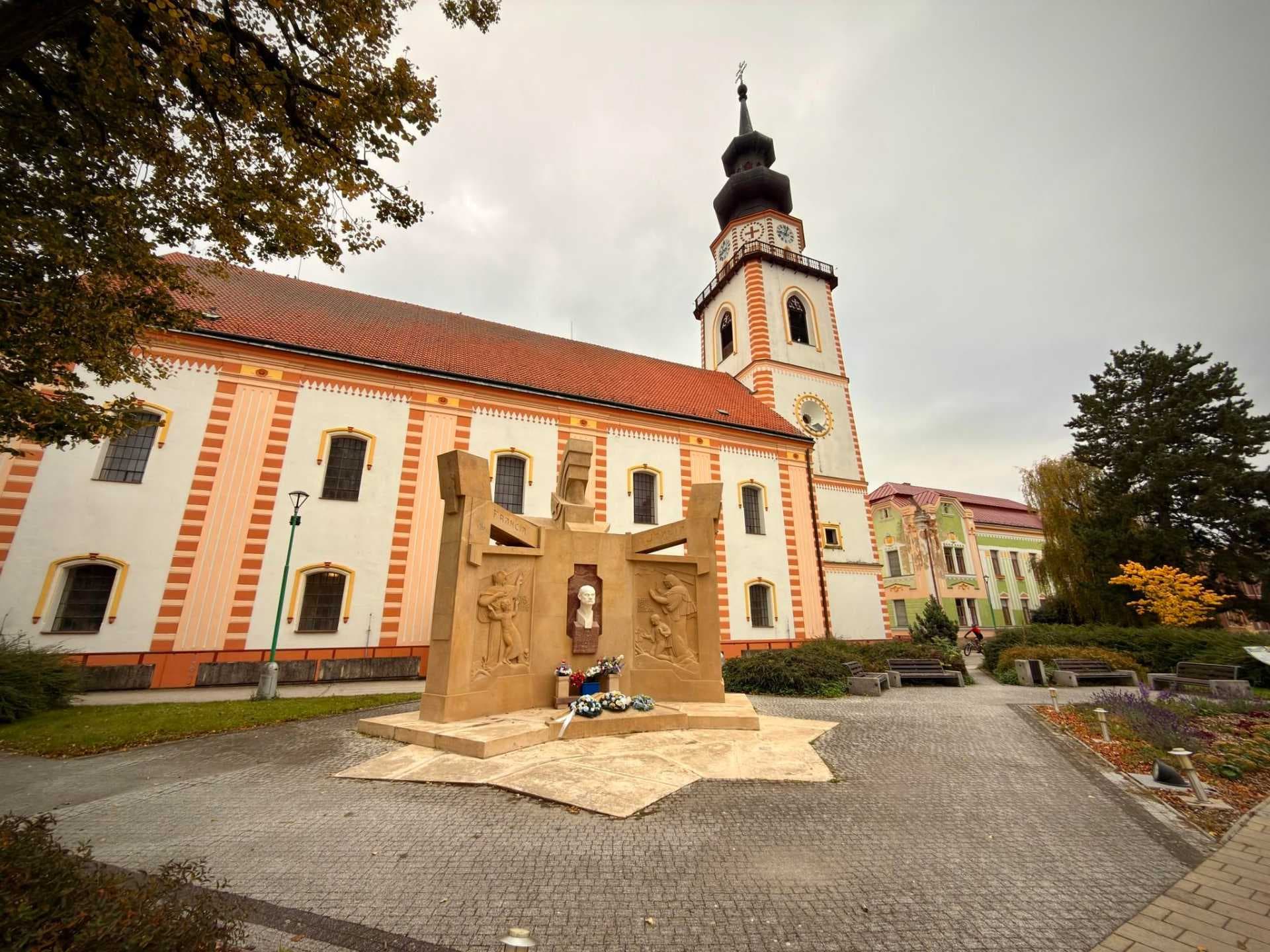 Historic church exterior with a striped facade and tall bell tower, with a stone memorial monument in the paved town square of Bánovce nad Bebravou.
