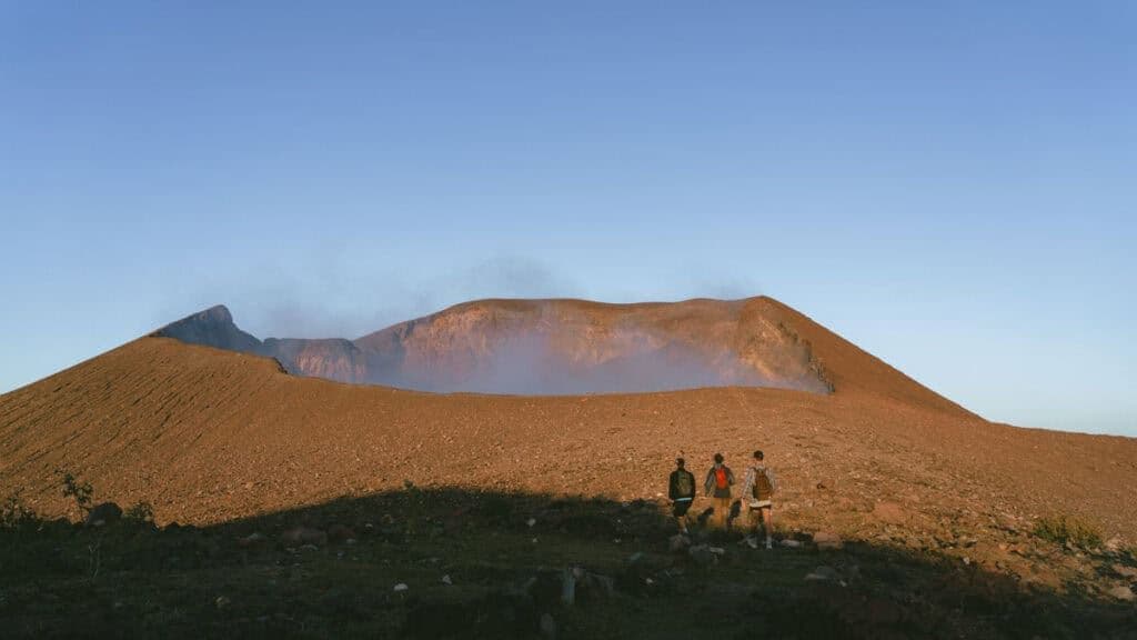 Volcanic landscape with a smoking crater, barren slopes, and three small hikers in the foreground under a clear sky.