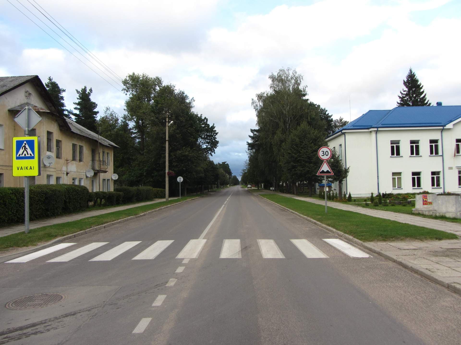 A quiet street scene in a Lithuanian town featuring a pedestrian crossing, a yellow sign reading Vaikai, and a white building with a blue roof.
