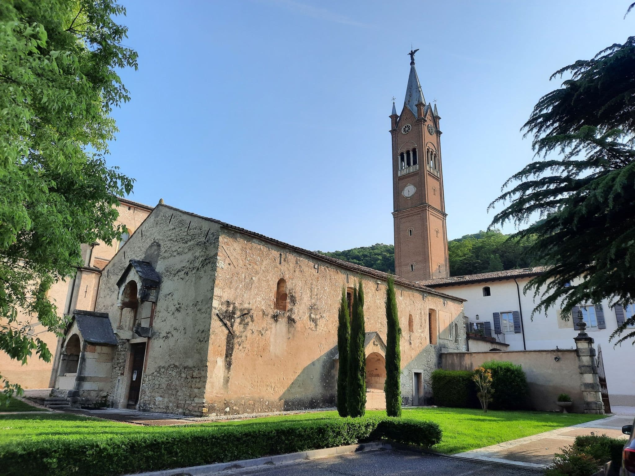 Exterior view of the historic Santa Maria church in Arbizzano, featuring a tall brick bell tower, weathered stone walls, and surrounding greenery under a clear