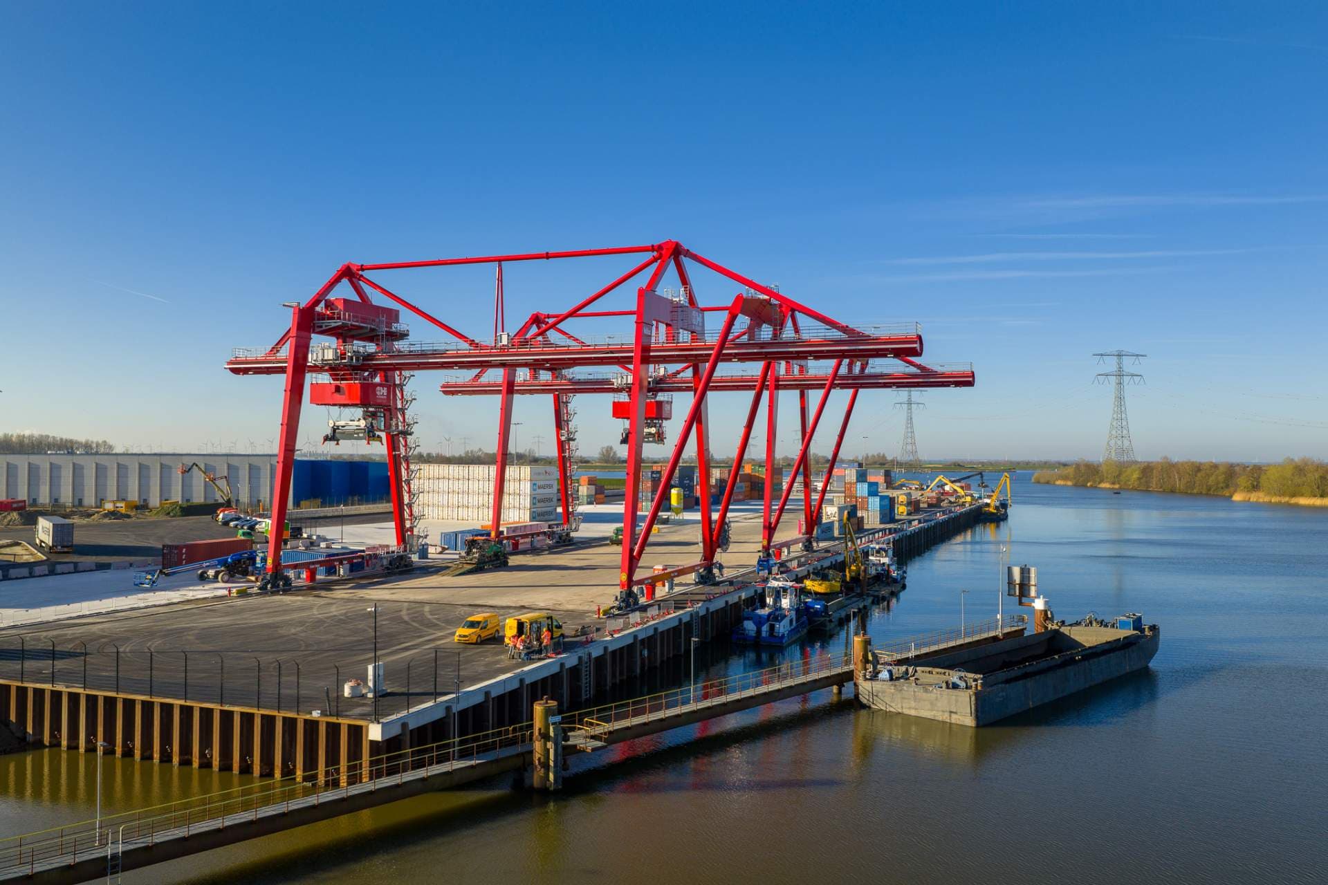 Aerial view of a modern industrial port featuring large red gantry cranes, shipping containers, and barges docked along a canal under a clear blue sky.