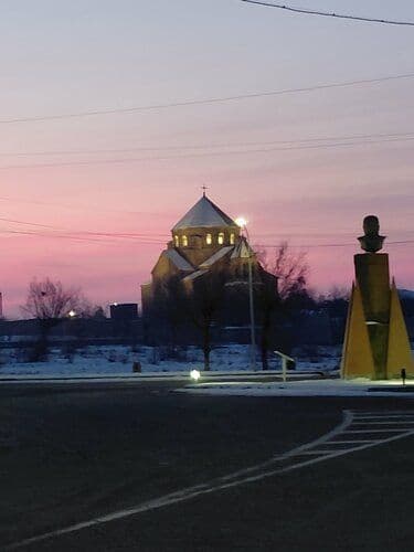 Dusk view of a church and monument in a snowy rural setting, with a road in the foreground.