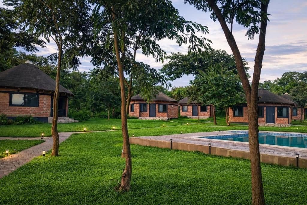 A real photograph of a resort or lodge complex with thatched-roof brick cottages, manicured lawns, trees, and a swimming pool near Lubambe, close to Chililabomb