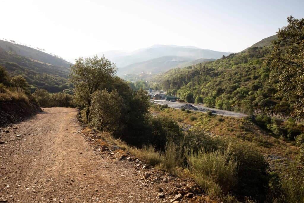 Dirt road winding through hilly, scrub-covered countryside with distant mountains and a small settlement visible in the valley.