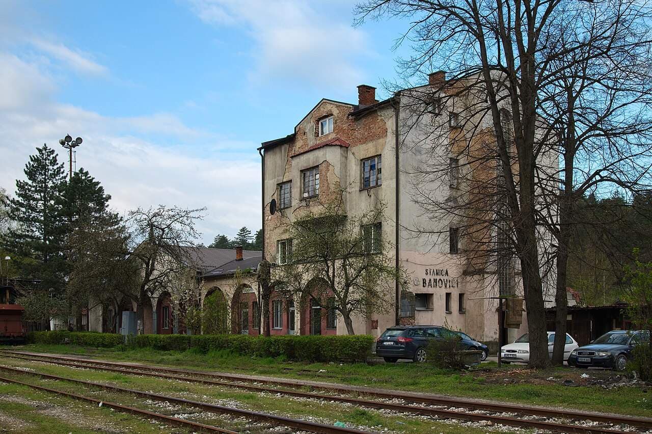 Exterior view of the Banovići railway station building with visible signage, adjacent train tracks, and parked cars under a blue sky.