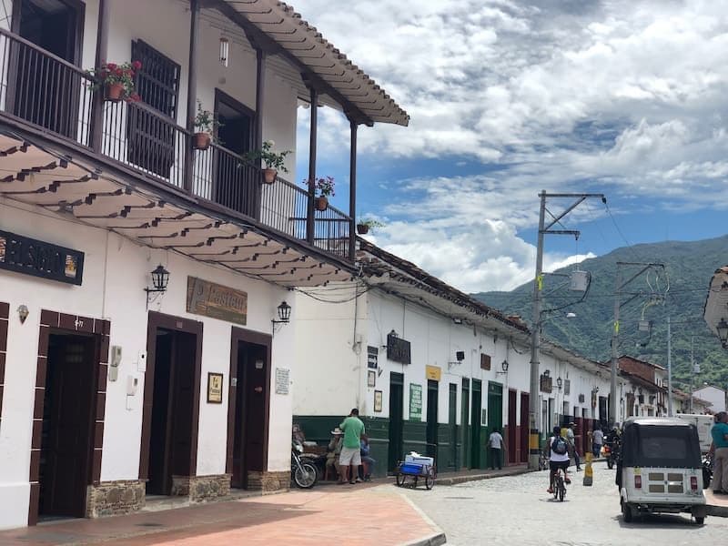 Street scene in Liborina with white colonial buildings, balconies, pedestrians, a cyclist, and a vehicle under a cloudy sky.