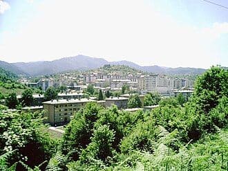 A distant landscape view of a town nestled in a valley with mountains in the background, featuring apartment blocks and lush green vegetation.