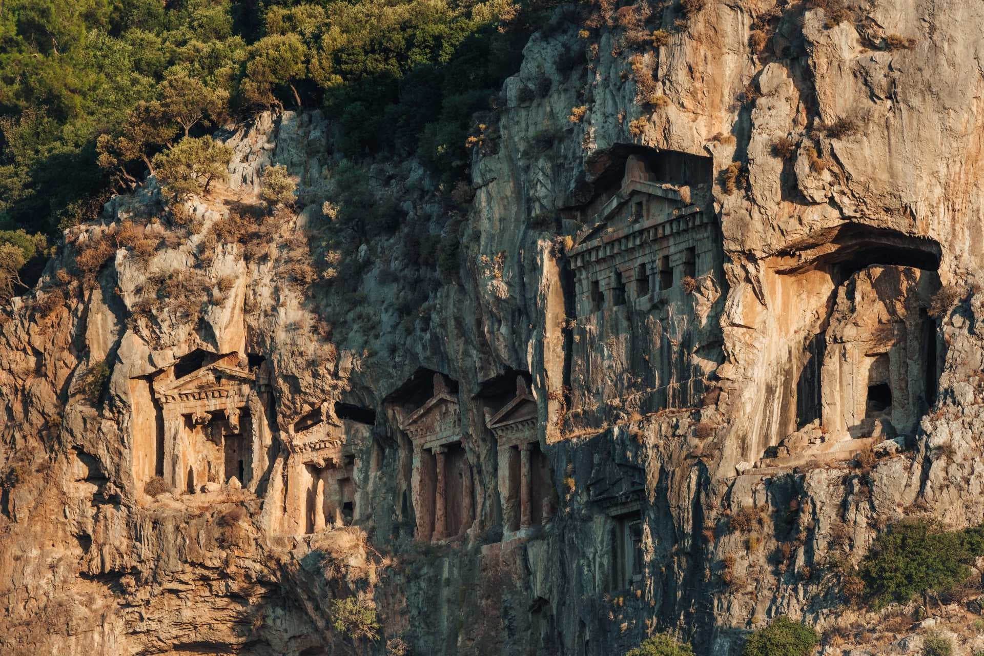 A row of ancient Lycian rock-cut tombs carved into a steep cliff face, featuring temple-style facades with columns and pediments.