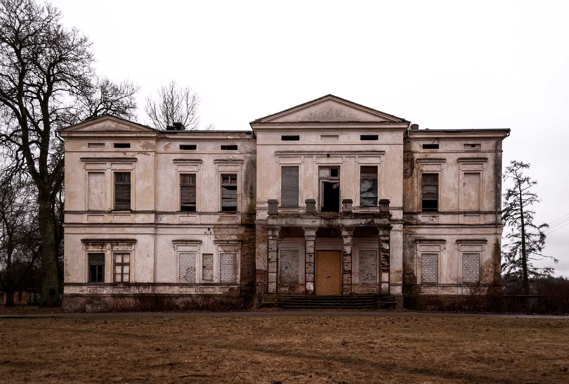 A wide shot of a dilapidated, classical-style manor house with boarded windows and peeling paint, set against a bleak, overcast sky.