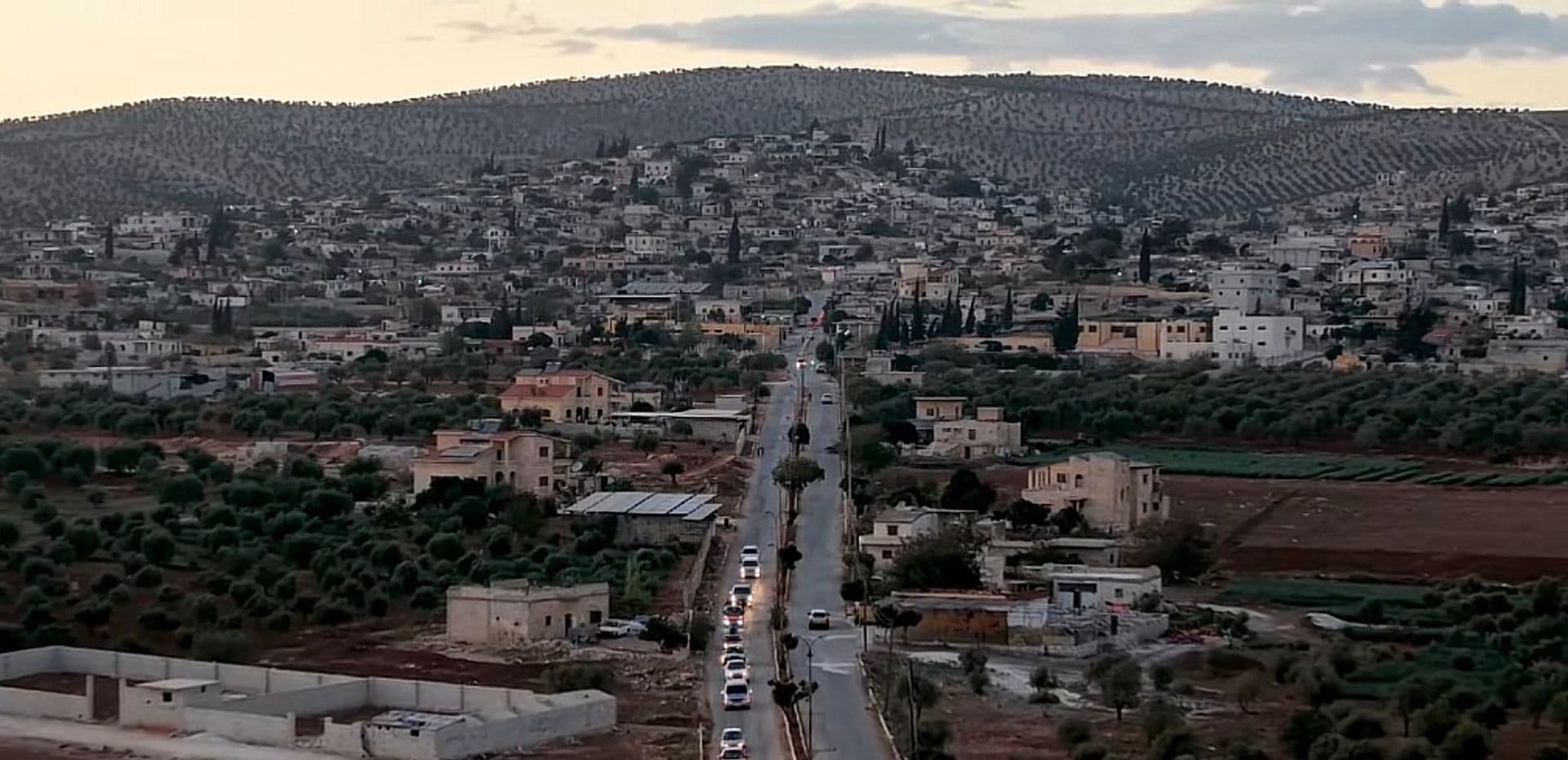 Wide aerial view of a hillside settlement with a central road, surrounded by olive groves and forested hills in the central Israel region.