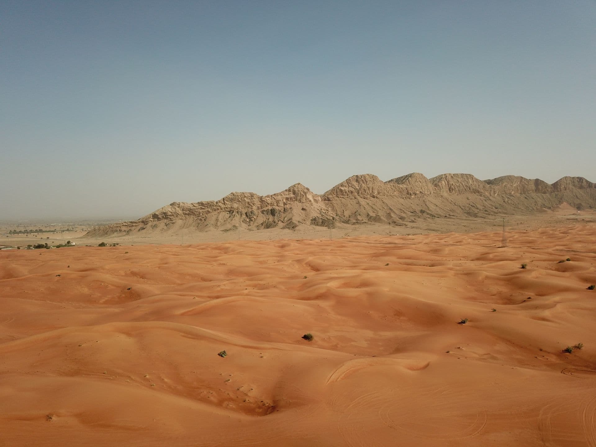 Vast orange sand dunes in the foreground with the rugged, rocky Al Faya mountain range rising in the background under a clear blue sky.