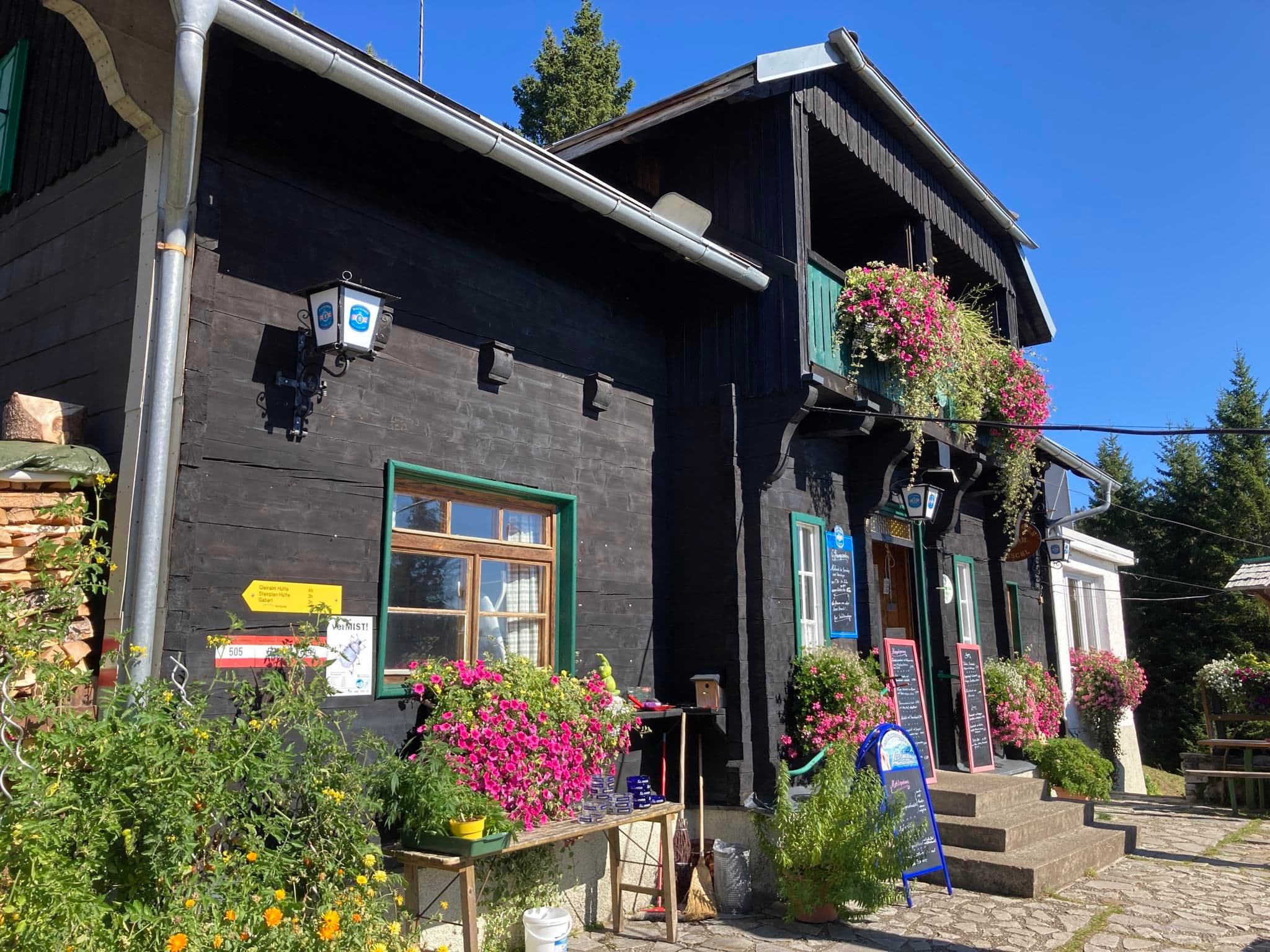 A traditional dark wooden building with vibrant pink flowers and green trim stands on a sunny day in a quiet village setting.