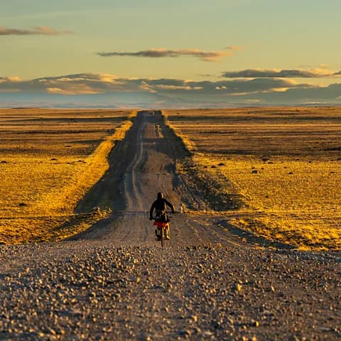 A cyclist rides an unpaved road through open pampa steppe under low autumn light with long shadows.