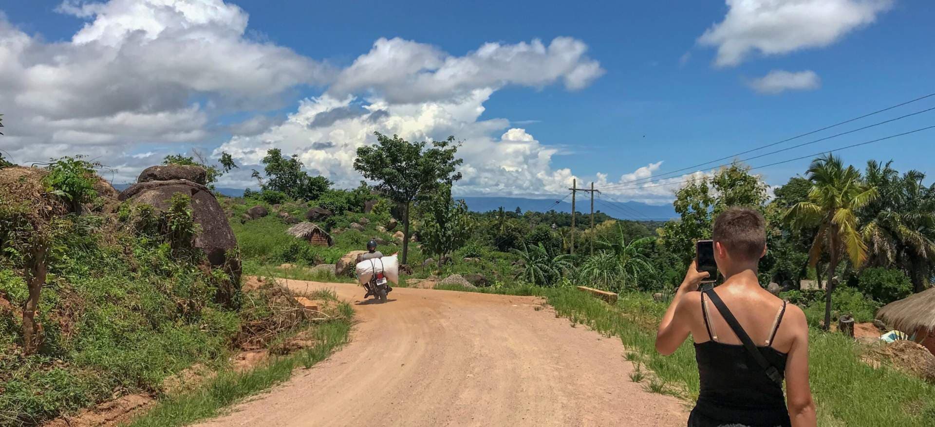 Wide rural scene with a dirt road, boulders, thatched huts, lush vegetation, and a motorbike in the Kitangari area of southern Tanzania.