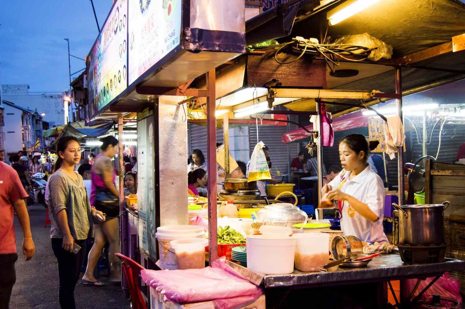 Nighttime street food stall with vendors preparing dishes, customers browsing, and illuminated signage in a busy market setting.
