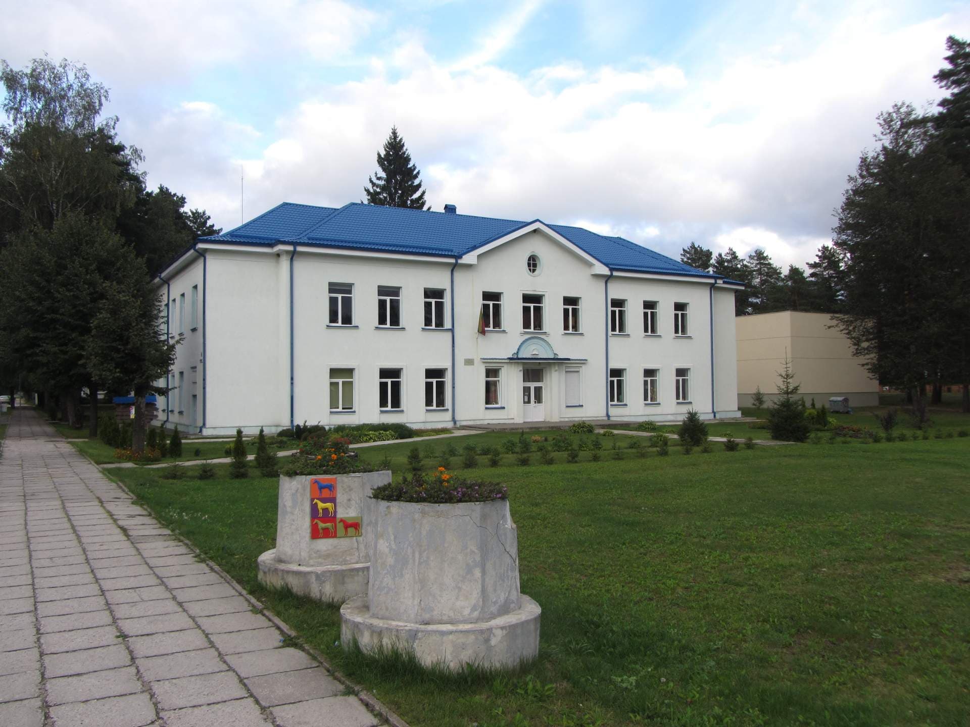 A white two-story building with a blue roof stands in a grassy area with trees and a paved walkway, representing the Baltoji Vokė area.