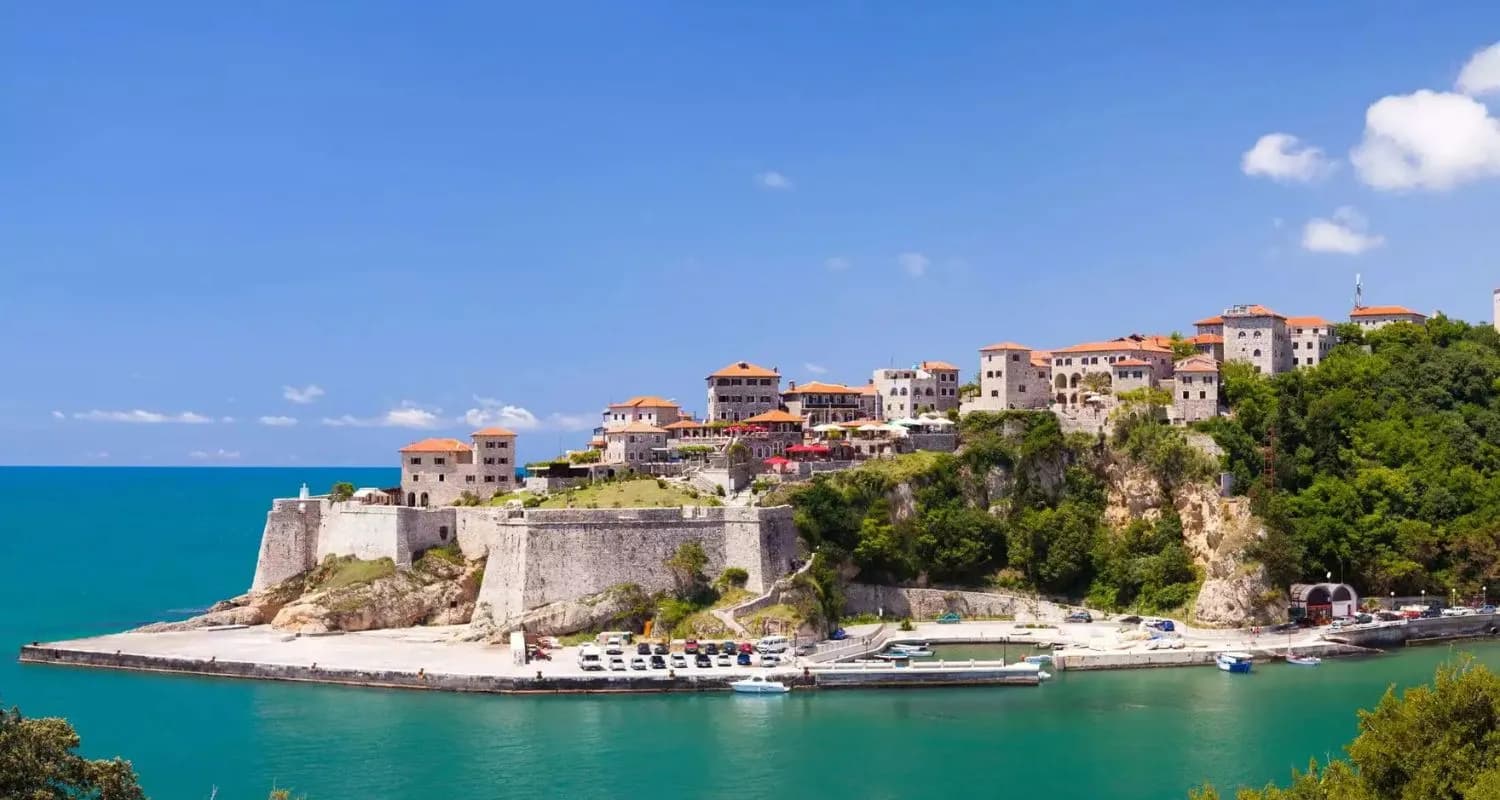 Wide exterior view of a historic coastal fortress town with stone walls, terracotta-roofed houses, and a harbor under a blue sky.