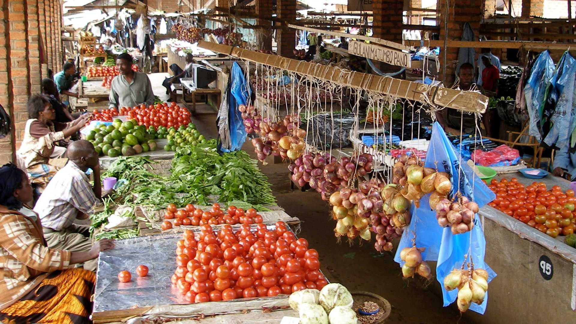Open-air market with vendors, fresh tomatoes, limes, leafy greens, and hanging onion bundles under a brick shelter.