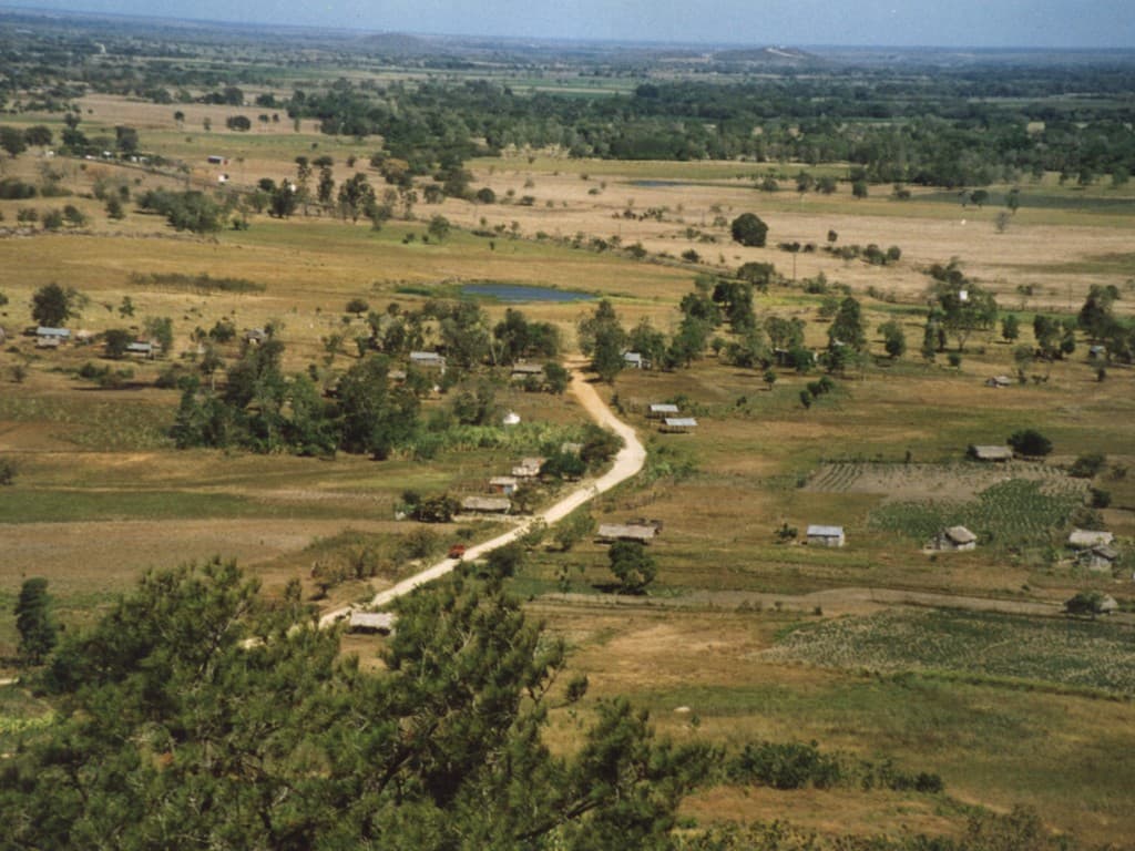 Aerial view of a rural landscape in Dajabon Province featuring a winding dirt road, scattered houses, and dry grassy fields.