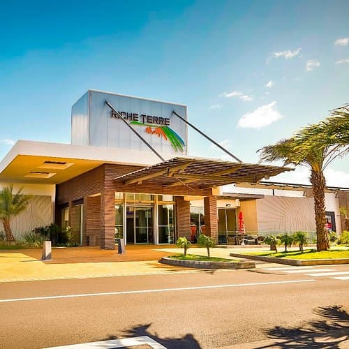 Exterior view of the Riche Terre shopping center entrance featuring modern architecture, a wooden canopy, and palm trees under a blue sky.