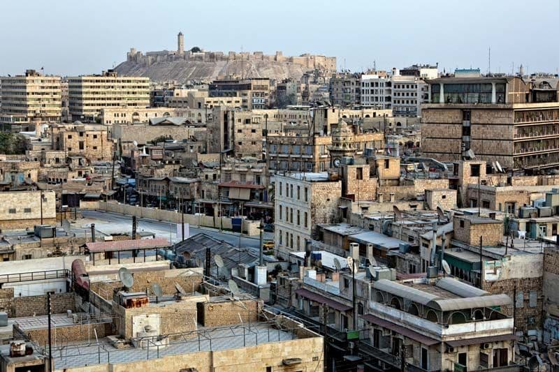 Aerial view of a dense historic town with stone buildings and a prominent hilltop fortress in the background.