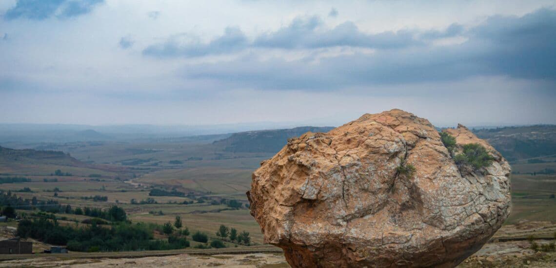Wide landscape photograph of a highland valley with rolling hills, scattered trees, and a large rock in the foreground under a cloudy sky.