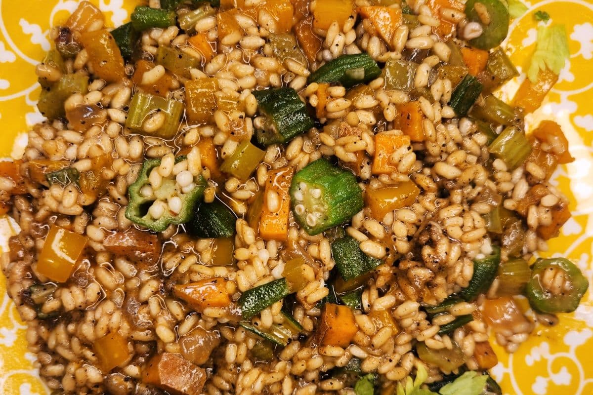 Close-up photograph of a cooked grain and vegetable dish with broth, okra, carrots, and peppers on a yellow patterned plate.