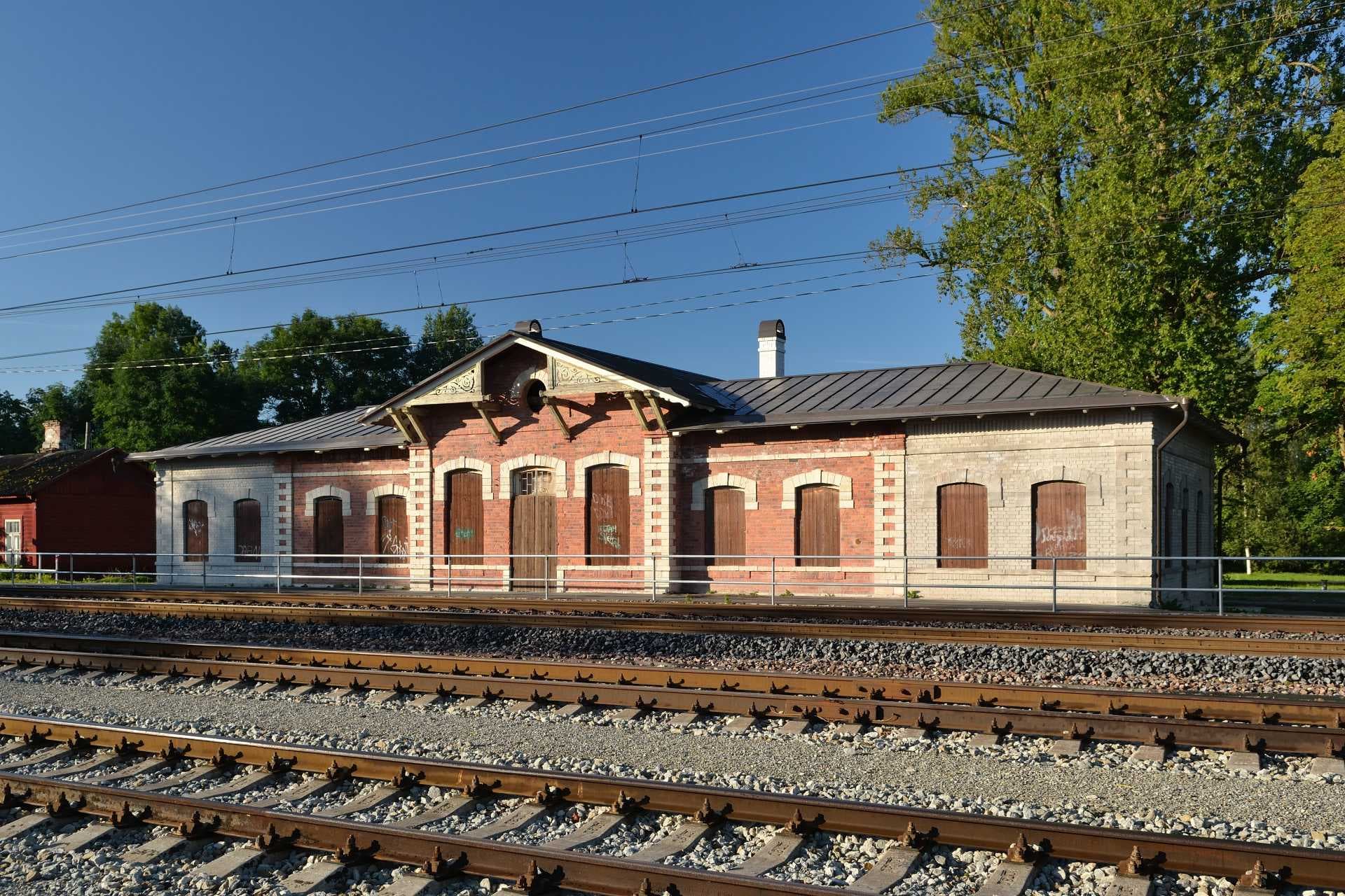 Exterior view of the historic Kehra Railway Station building with red brick and white stone facade, situated beside multiple railway tracks under a clear blue s