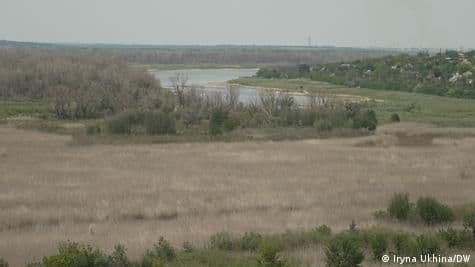 Wide landscape view of the Kachovka Reservoir and Dnipro shoreline near Dniprorudne with dry grasslands and sparse trees under an overcast sky.