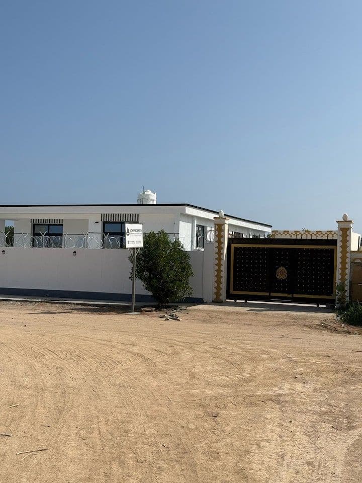 White single-story guesthouse exterior with black-and-gold gate, barbed wire, and a sign, set on a dirt lot under a clear blue sky.