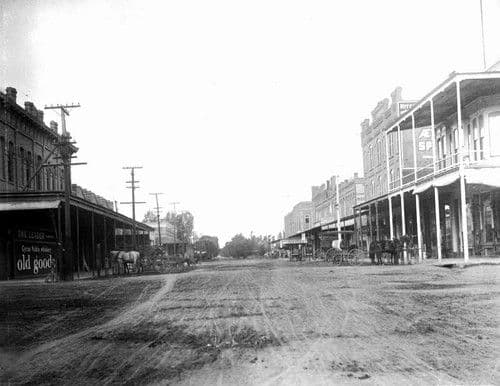 Historic black-and-white photograph of a wide downtown street with storefronts, buildings, and horse-drawn carriages.