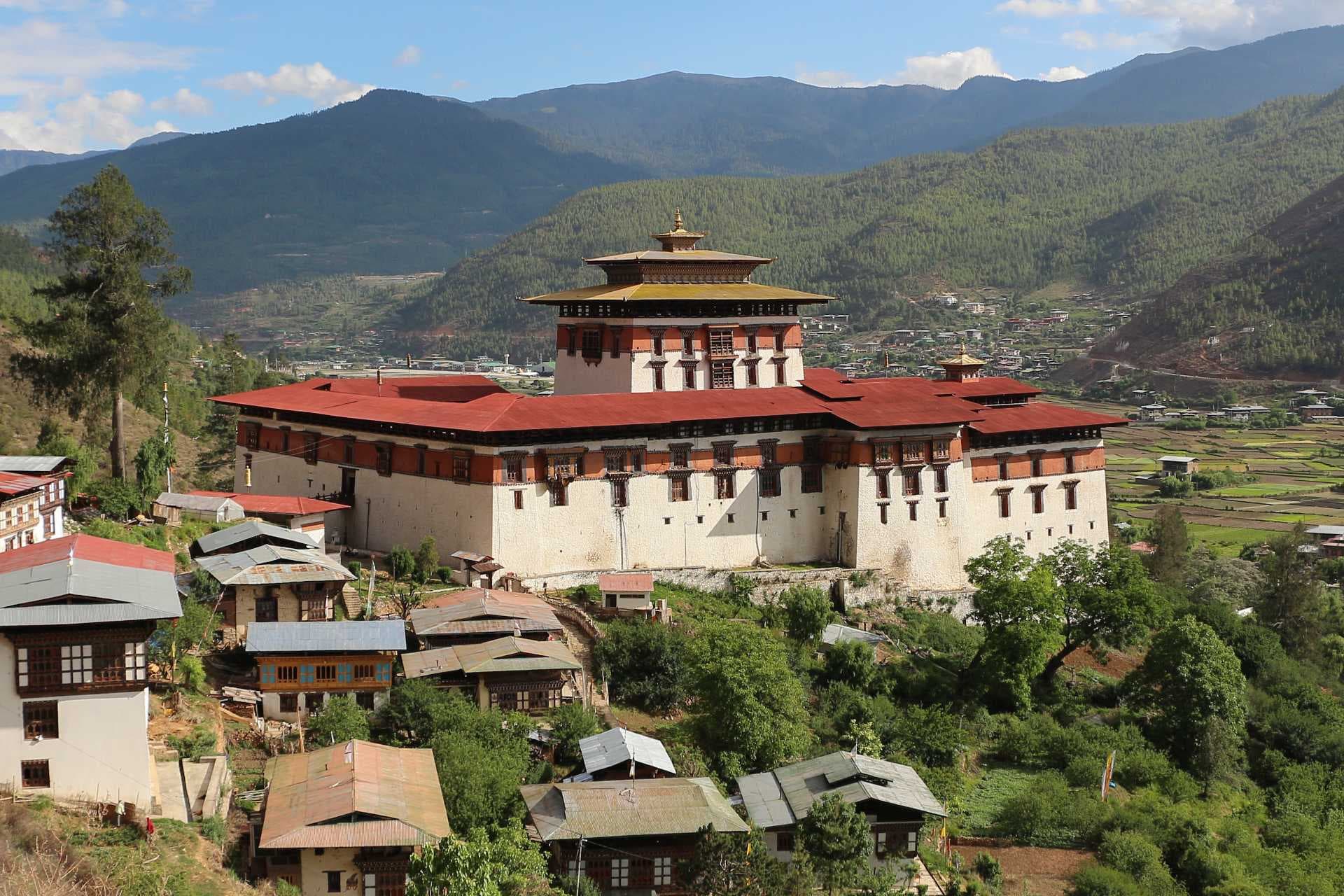 Wide exterior view of Paro Dzong fortress-monastery in a mountain valley, with surrounding village houses and forested hills.