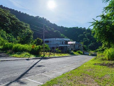 A paved rural road winds through a lush green landscape in Saint Andrew, Jamaica, with a building nestled against a forested hillside under a bright sun.